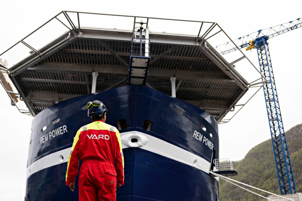 En VARD ansatt står i rød dress foran et blått skip.