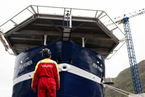En VARD ansatt står i rød dress foran et blått skip.