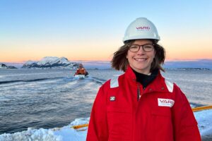 Ei ung dame med brunt hår, briller, rød VARD drakt og hvit vard hjelm, foran en vard båt som kjører ut mot havet i et vinterlandskap.